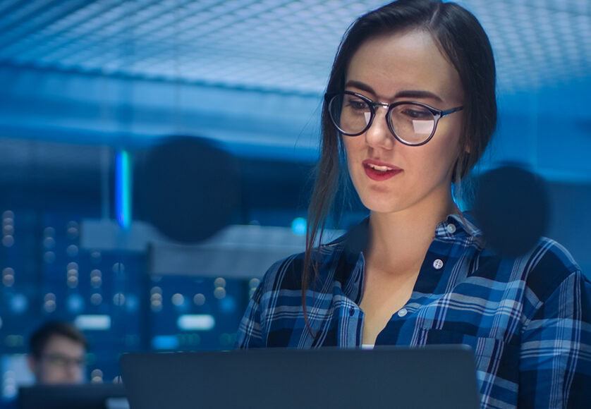 Portrait of a Smart Seductive Young Woman Wearing Glasses Holds Laptop. In the Background Technical Department Office with Specialists Working and Functional Data Server Racks