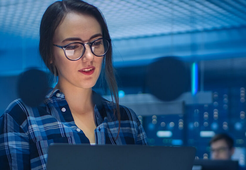 Portrait of a Smart Seductive Young Woman Wearing Glasses Holds Laptop. In the Background Technical Department Office with Specialists Working and Functional Data Server Racks