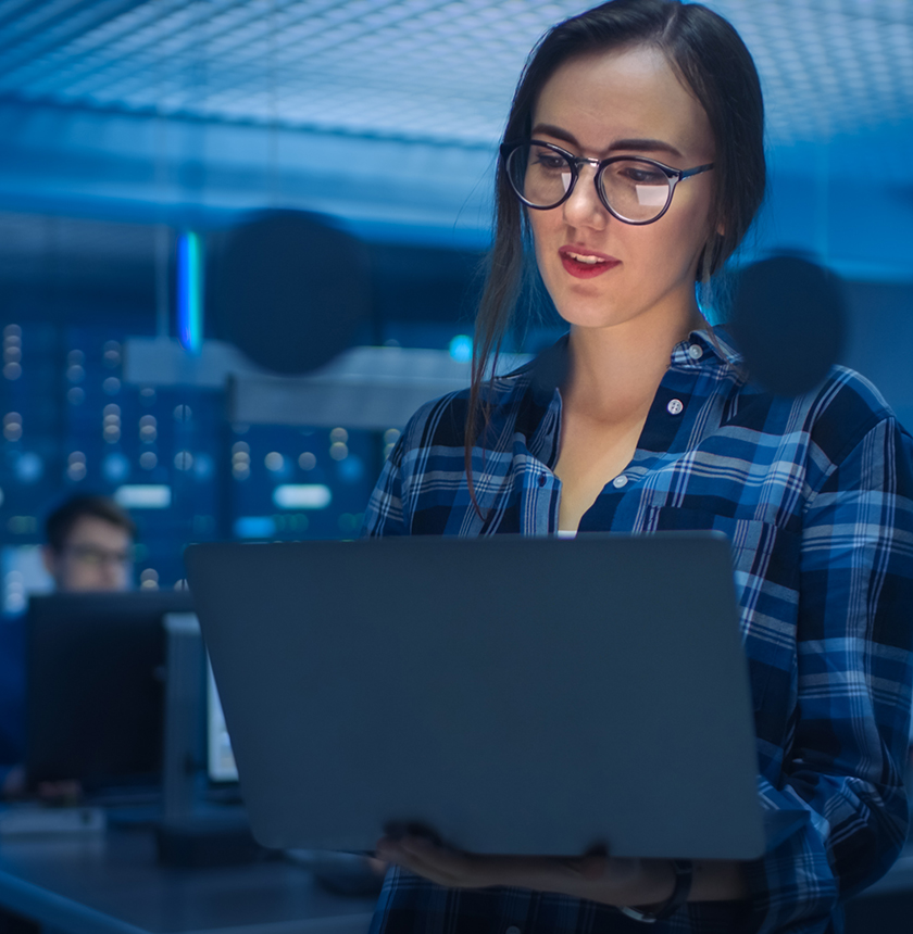 Portrait of a Smart Seductive Young Woman Wearing Glasses Holds Laptop. In the Background Technical Department Office with Specialists Working and Functional Data Server Racks