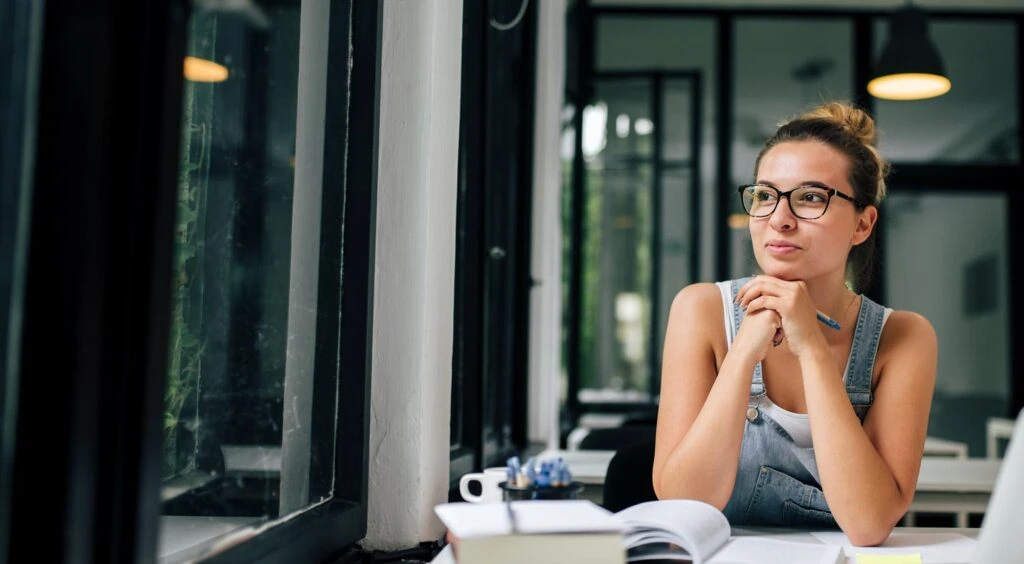 Young woman studying at a desk with large windows in the background.